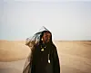 Dua Saleh is standing in the desert, wearing all-black and heavy golden jewelry, with a solemn expression.