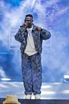Onesimus, dressed in a patterned outfit, sings on a blue-lit stage with smoke behind him.