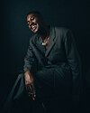 Ghanaian musician RCee, smiling, dressed in a dark suit, seated against a black background, lit with dramatic side lighting.