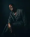 Ghanaian musician RCee, smiling, dressed in a dark suit, seated against a black background, lit with dramatic side lighting.