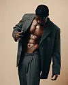 Ghanaian musician RCee, wearing an oversized dark suit, stands against a plain beige background with his jacket open.