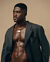 Ghanaian musician RCee, in an open dark blazer and chain necklace, stands against a beige backdrop.