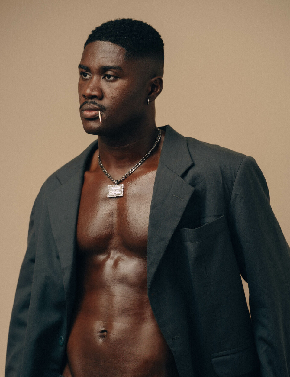 Ghanaian musician RCee, in an open dark blazer and chain necklace, stands against a beige backdrop.