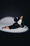 A woman wearing a black suit and red lipstick is sitting on the floor of what appears to be a photo studio.