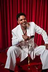Gil Semedo, in a white suit, is sitting and smiling with a microphone in front of red curtains.