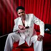 Gil Semedo, in a white suit, seated in front of red drapes, holding a microphone and a black prop.