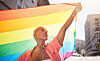 A Black woman with short blonde hair holding a rainbow flag overhead on a sunny urban street.
