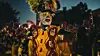 A masked performer known as a Kaapse Klopse minstrel in a gold outfit and feathered headdress, at a nighttime festival.
