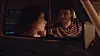 Two people inside a car at night, seen through the window with warm light and reflections.