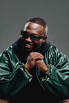 Man in sunglasses smiling with clasped hands, wearing a green jacket against a gray background.