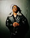 NO11, wearing glasses and a black leather jacket, stands against a textured wall with hands clasped.
