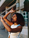 Close-up portrait of Meron T outdoors in a white sleeveless top, raising her arms near her face.