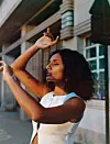 Close-up portrait of Meron T outdoors in a white sleeveless top, raising her arms near her face.