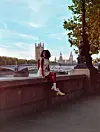 Meron T sits on a stone wall by the River Thames in London, wearing a white outfit, with the city visible in the background.
