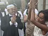 A man wearing a black suit and a Sudanese hat and a woman in a white wedding dress are smiling and dancing in a garden.