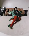Shekhinah IS seated on a rug in a minimalist living room with a brown couch and audio equipment. She leans against a brown sofa and wears an oversized green t-shirt, red stockings, and green shoes.