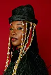 Portrait of Fatoumata Diawara in a black hat with shell-adorned braids on a red background.