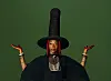 Fatoumata Diawara, in a large black hat and dark outfit, poses with raised hands against a green background.