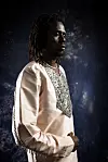 Emmanuel Jal, in a cream embroidered tunic, poses in profile against a dark background.