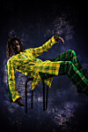Emmanuel Jal, reclining on a chair in a yellow and green plaid outfit against a dark background.