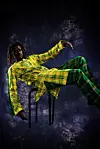 Emmanuel Jal, reclining on a chair in a yellow and green plaid outfit against a dark background.