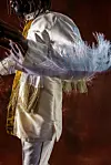 Emmanuel Jal is wearing white ceremonial attire with feathered adornments against a dark backdrop.