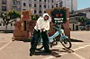 Person in a white hoodie stands next to a light blue custom scooter in a parking lot with boxes and graffiti art.
