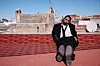 Man seated on a rooftop with buildings, antennas, and a satellite dish in the background.
