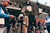 Crowd of people gathered under an overpass near weathered buildings and temporary structures.