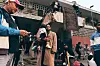 Crowd of people gathered under an overpass near weathered buildings and temporary structures.
