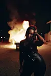 Nikita Kering’, in a black leather outfit, poses at night in front of a burning car.