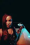 Nikita Kering’ poses at night beside a car with a damaged windshield, holding a bat and wearing a black leather outfit.