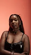 Studio portrait of Nikita Kering’ in a black shiny top against a coral background.