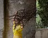 Neta Elkayam is wearing a yellow dress that is half made of plastic and carrying a bunch of wooden sticks over her head.