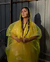 Neta Elkayam is wearing a yellow dress under a plastic overall, her hair is braided and she is looking into the distance.