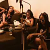 Three women sit on a couch in a warm-toned room, using phones near a microphone stand and camera setup.