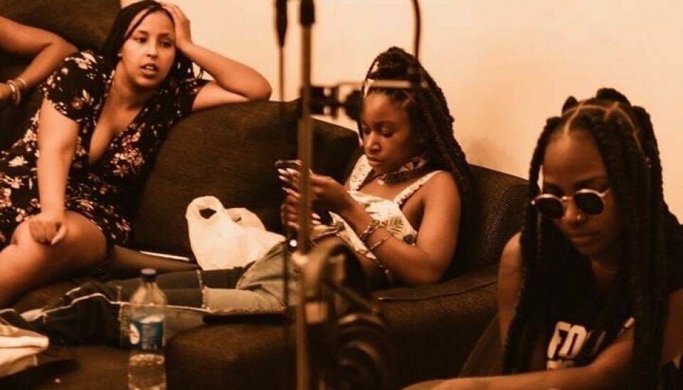 Three women sit on a couch in a warm-toned room, using phones near a microphone stand and camera setup.