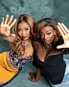 Two women, duo We are Nubia, pose close to the camera in a studio setting, hands raised and wearing stylish outfits.