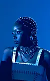 Blue-lit portrait of Karun wearing a studded headpiece and layered jewelry.