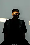 Vanco, wearing glasses and dark clothing, is standing in shadow against a plain wall with a strip of sunlight.