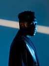 Profile portrait of Vanco in a dark jacket against a blue background with a diagonal light beam.