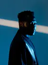 Profile portrait of Vanco in a dark jacket against a blue background with a diagonal light beam.