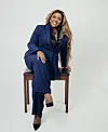Portrait of Gabrielle Chams seated on a chair in a navy blue suit against a white background.