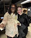 Whoopi Goldberg and Leleti Khumalo are smiling together in a backstage room with mirrors and equipment behind them.