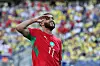 Morocco player Ismael Saibari, in a red jersey, salutes after scoring in front of a blurred crowd.