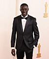 Khaby Lame stands in a black tuxedo at the Academy Awards red carpet backdrop.