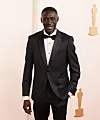 Khaby Lame stands in a black tuxedo at the Academy Awards red carpet backdrop.