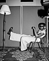 Black-and-white image of a person seated in a folding chair next to a floor lamp and studio gear.