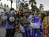 Two smiling men are holding large castanets and playing them, wearing colorful blue-and-green traditional Moroccan attire.