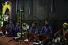 Seven men are on cushions on the floor, singing into microphones and playing the qraqeb. In the middle is a man wearing different clothes, playing the guembri.